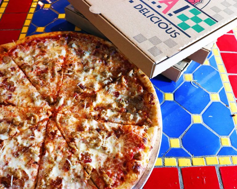 Close-up of a fresh pizza topped with sausage and cheese next to stacked takeout boxes on a colorful tiled table.