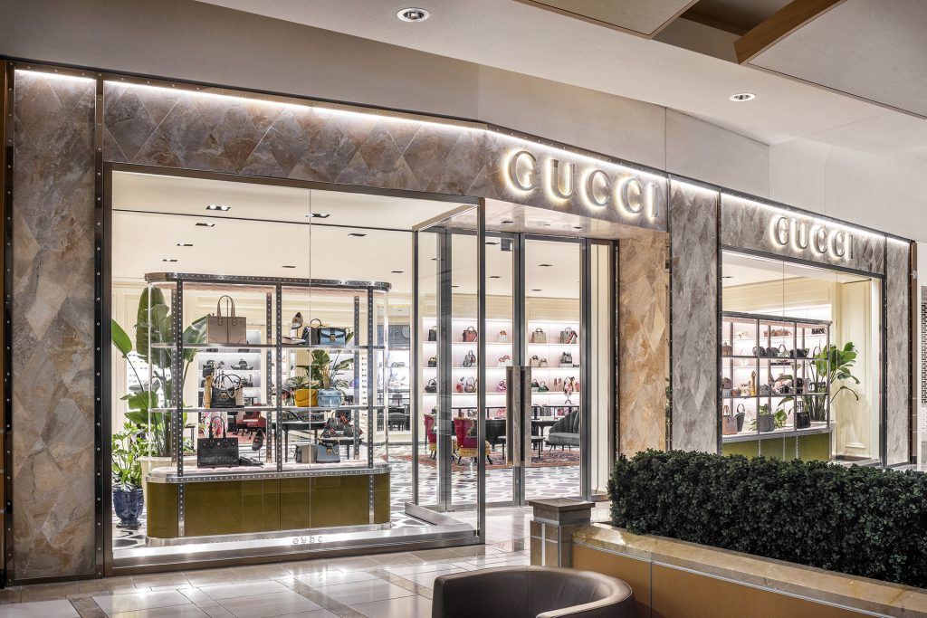 Exterior entrance of a Gucci store in a shopping mall with glass doors and illuminated signage.