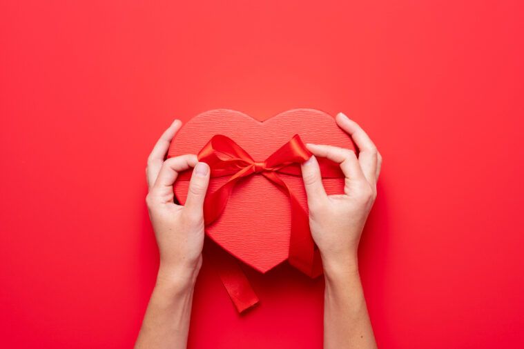Close up on female hands holding a gift in a pink heart presents for valentine day, birthday, mother's day. Flat lay. Symbol of love. Valentines day background with a gift boxes on concrete board.