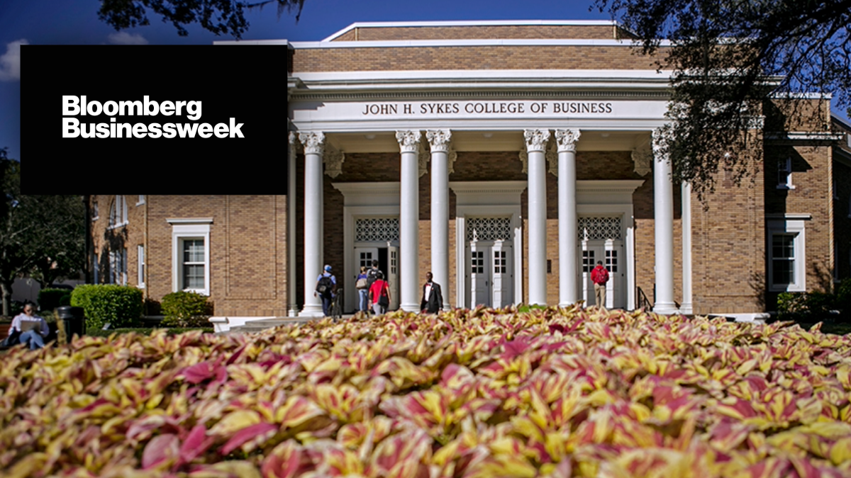 Front entrance of the University of Tampa’s John H. Sykes College of Business with students walking across campus, overlaid with the Bloomberg Businessweek logo.