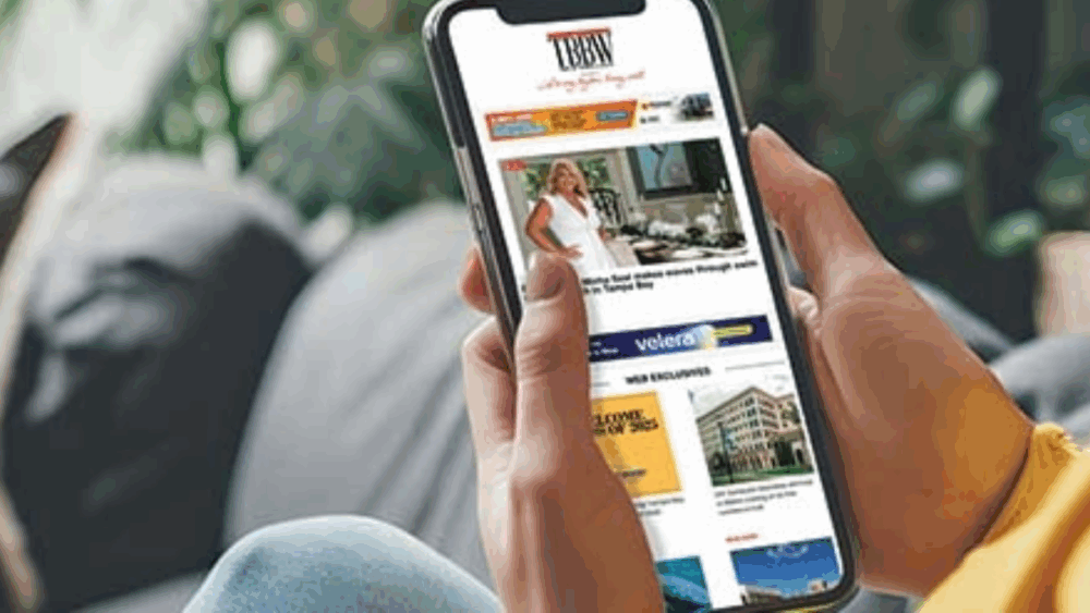 A person holds a smartphone displaying the Tampa Bay Business & Wealth website, showing local business headlines and feature stories on the screen.