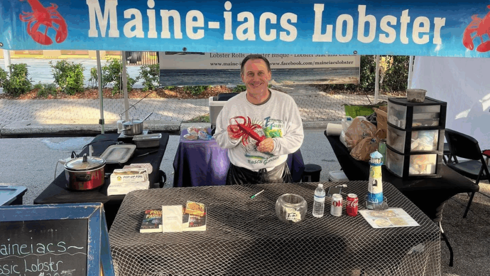 Guy Cote of Maine-iacs Lobster stands behind his booth at a Tampa Bay Markets event, smiling while holding a red lobster prop, with a banner reading “Maine-iacs Lobster” displayed above him.