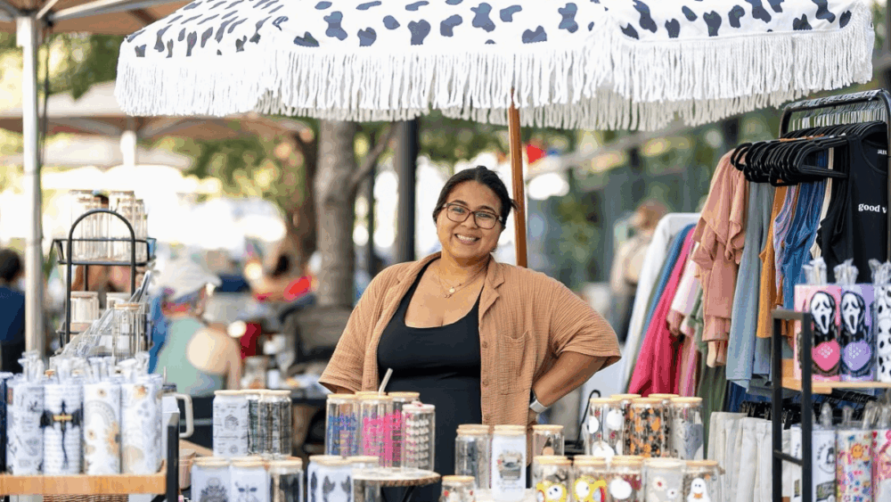 A smiling vendor stands under a fringed umbrella surrounded by handmade tumblers and clothing racks at the Fresh Market at Hyde Park Village, part of the Tampa Bay Markets network.