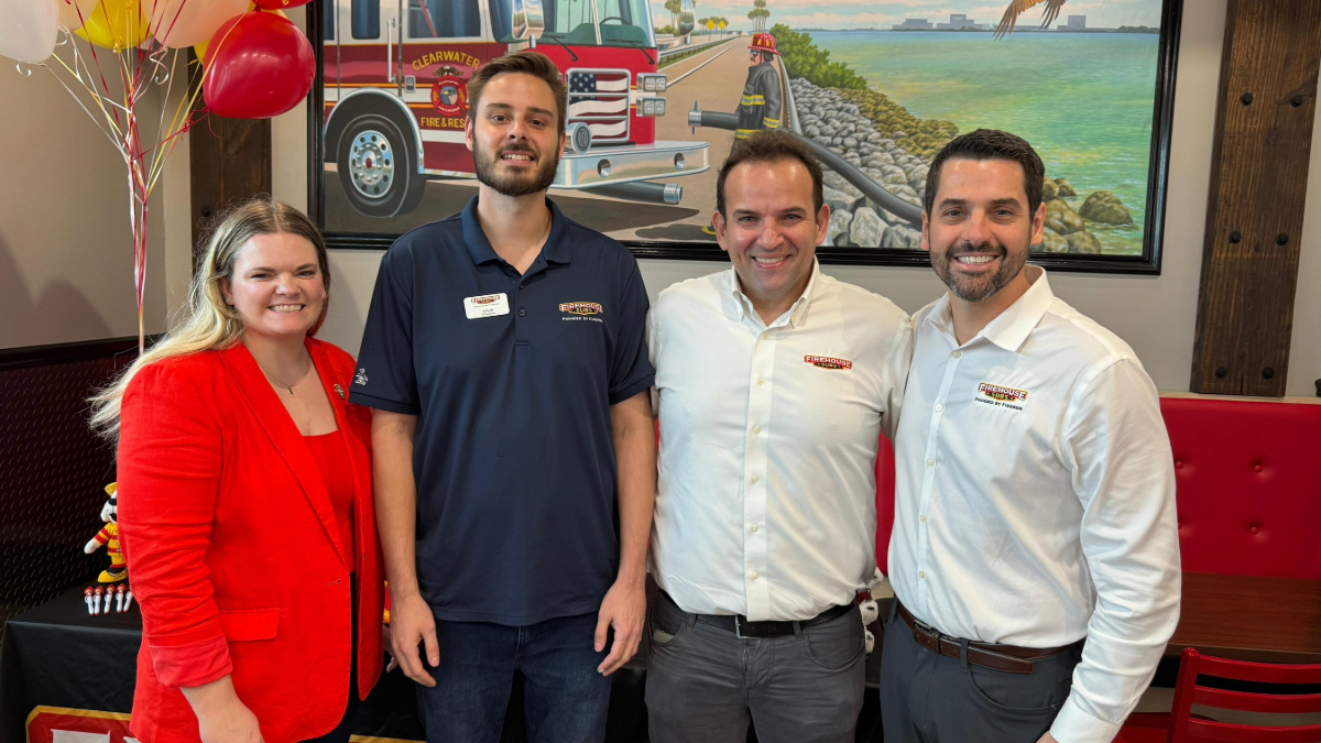 Four people stand together inside the new Firehouse Subs restaurant in Clearwater, smiling for a photo in front of a mural featuring a Clearwater Fire & Rescue truck.
