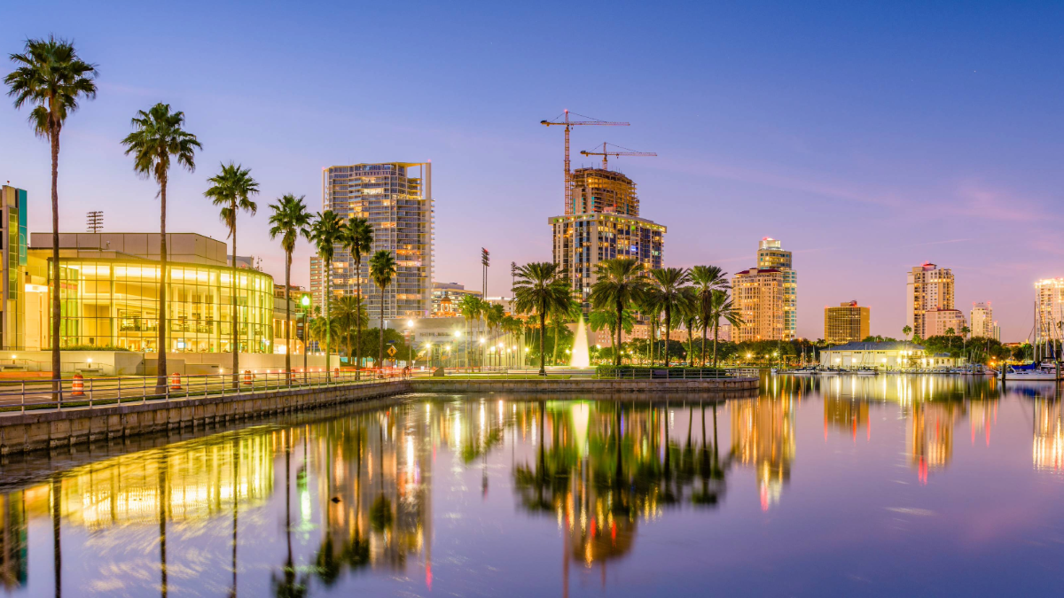 Downtown St. Petersburg skyline at sunset reflecting over the waterfront, showcasing high-rise buildings, palm trees, and construction cranes against a pink and purple sky.