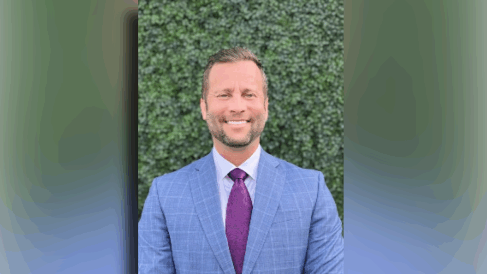 A man in a blue suit and purple tie smiles in front of a green wall.