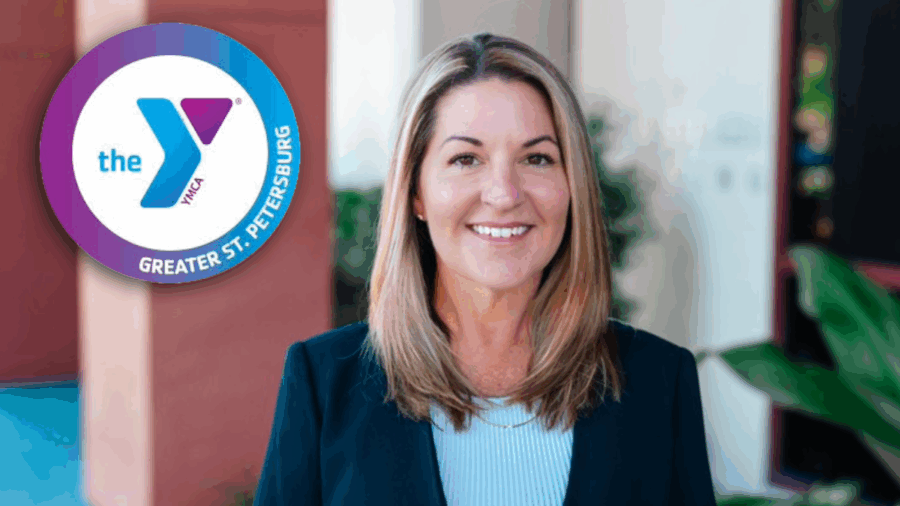 Nicci Bucher, newly appointed CEO of the YMCA of Greater St. Petersburg, stands outside a YMCA facility, smiling confidently. The YMCA logo is displayed beside her.