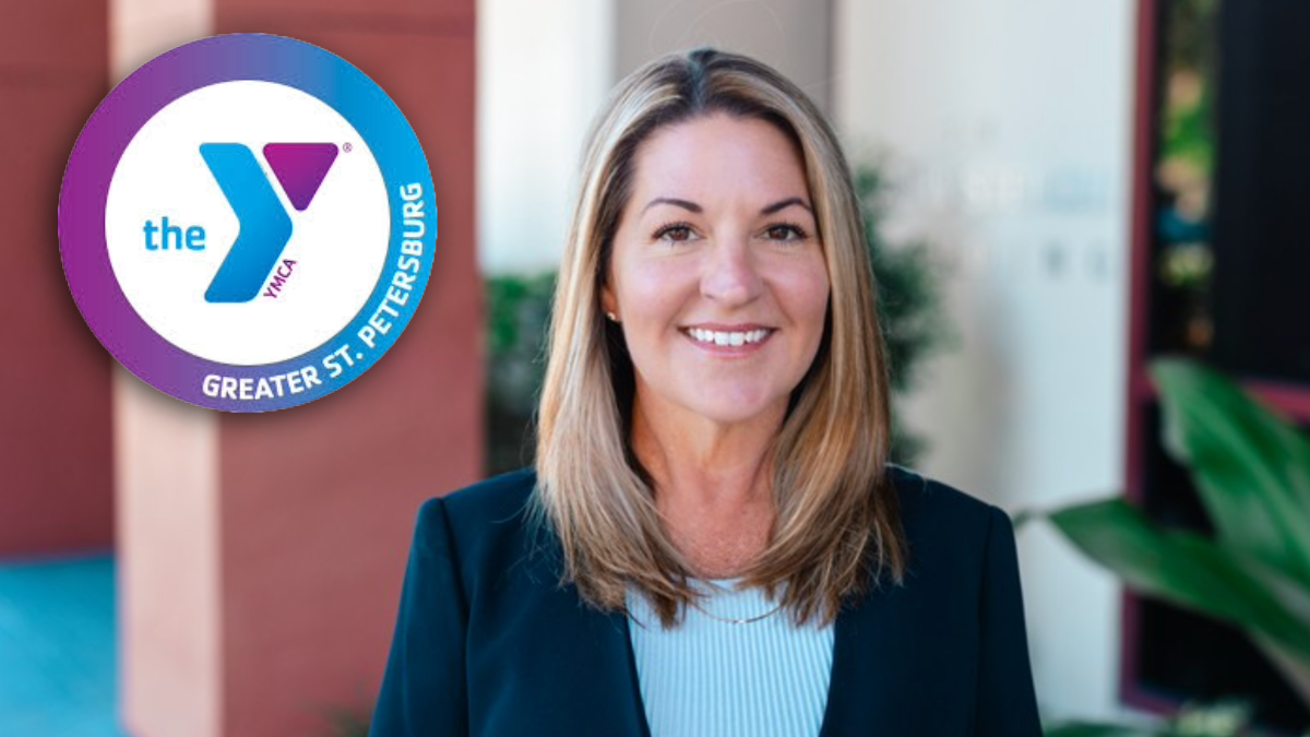 Nicci Bucher, newly appointed CEO of the YMCA of Greater St. Petersburg, stands outside a YMCA facility, smiling confidently. The YMCA logo is displayed beside her.