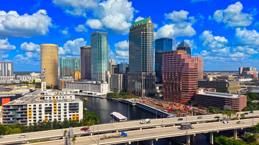 Downtown Tampa skyline with office towers and residential buildings along the Hillsborough River under a bright blue sky.