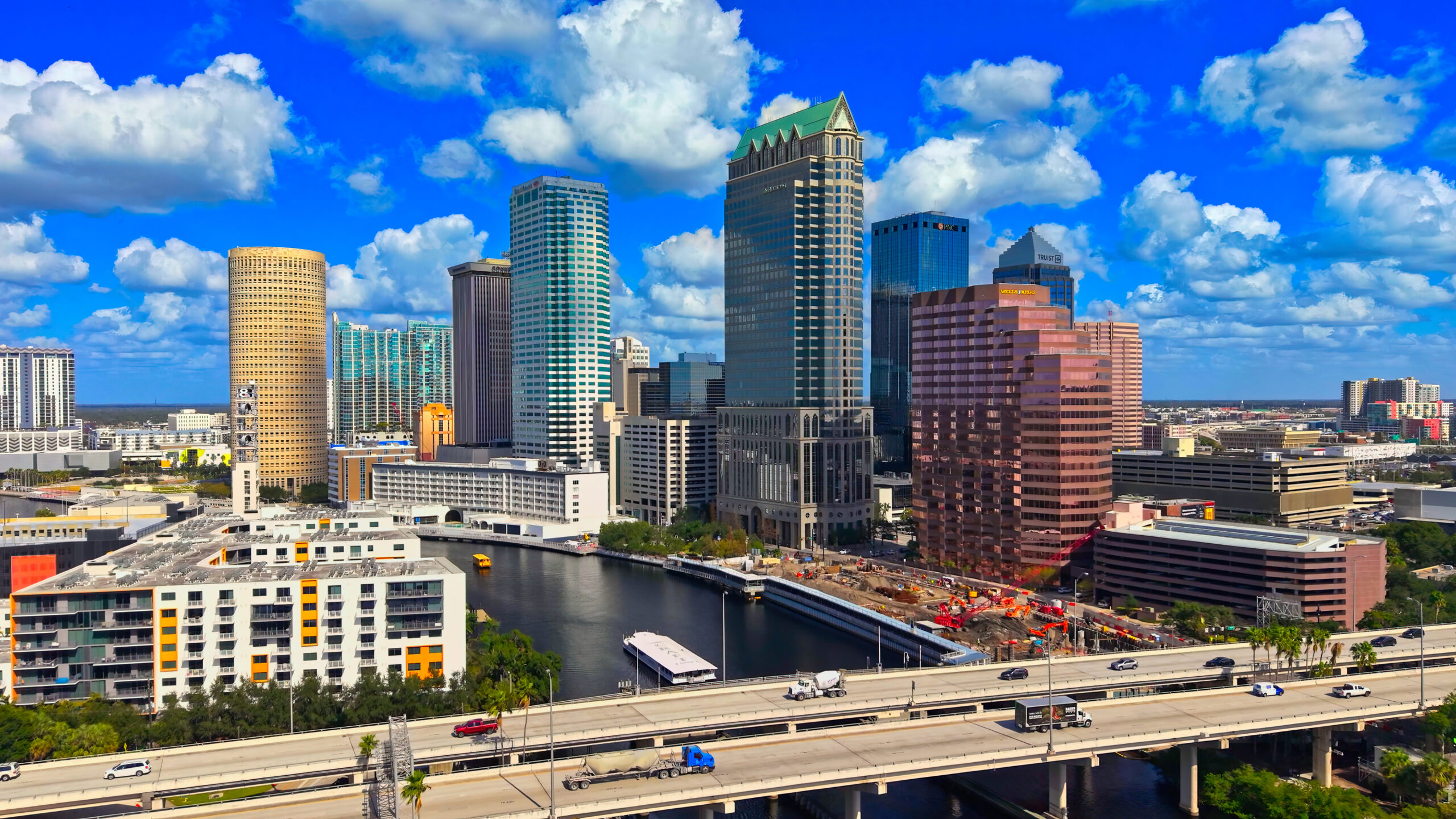 Downtown Tampa skyline with office towers and residential buildings along the Hillsborough River under a bright blue sky.