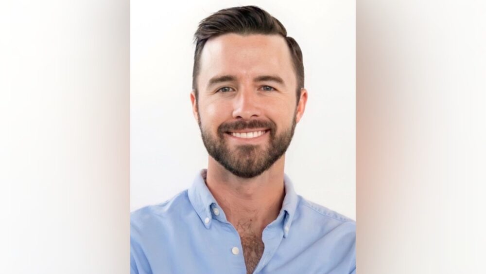 Riley Walker smiling in a light blue button-down shirt against a white background.