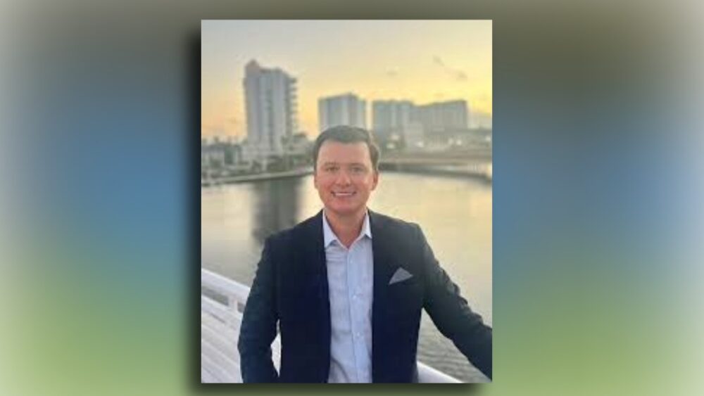 Will Freeman, a man in a navy suit, standing on a bridge overviewing downtown Tampa at sunset.