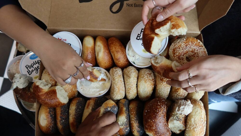 Hands grabbing fresh bagels from a PopUp Bagels box with assorted cream cheeses