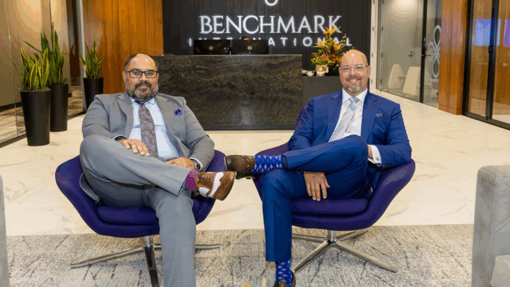 Benchmark International partners Dara Shareef and Jared Hardin seated in front of the company’s logo wall at its Tampa office, with Shareef on the left and Hardin on the right