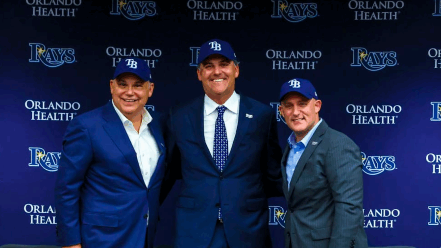 Three members of the Tampa Bay Rays’ new ownership group pose together wearing team hats at a press conference backdrop featuring the Rays and Orlando Health logos.