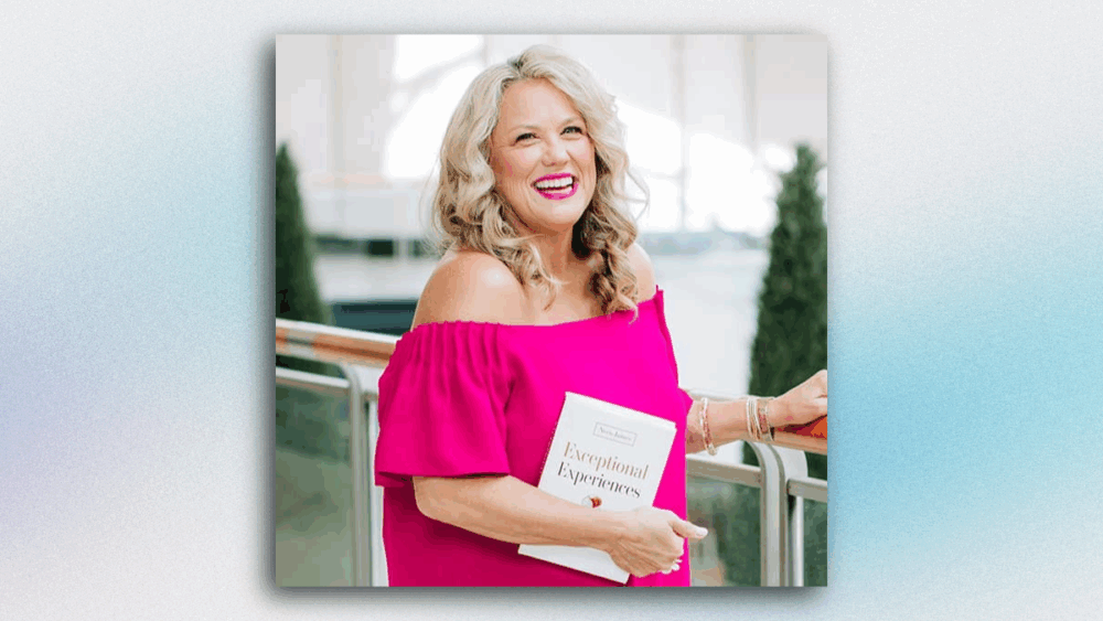 Neen James smiles while holding her book Exceptional Experiences, wearing a bright pink dress and standing along a balcony.