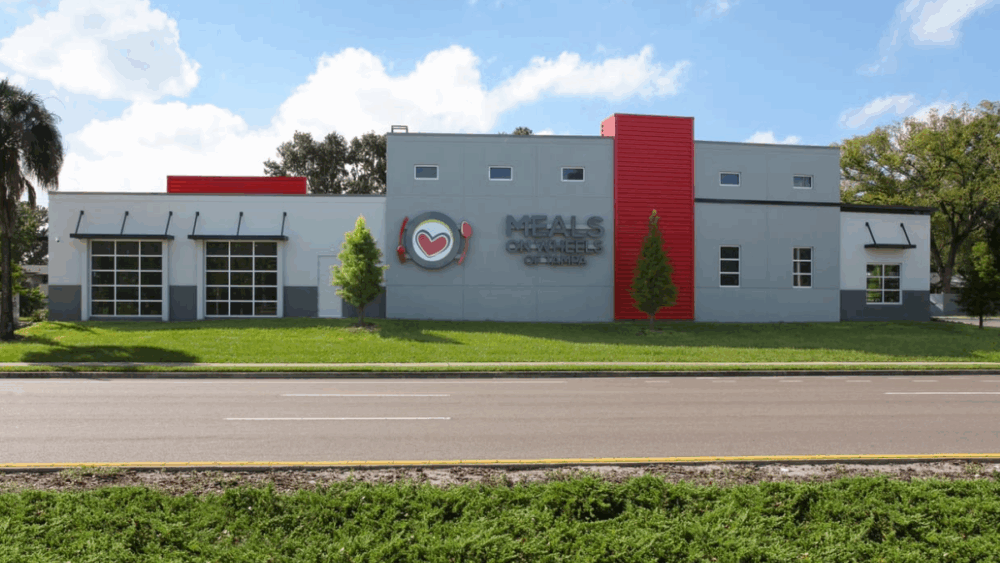 Meals On Wheels of Tampa headquarters building with a modern gray and red exterior, showcasing the nonprofit’s heart and utensils logo on the front facade.
