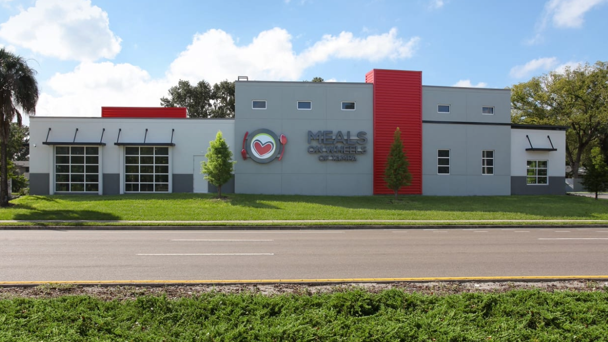 Meals On Wheels of Tampa headquarters building with a modern gray and red exterior, showcasing the nonprofit’s heart and utensils logo on the front facade.
