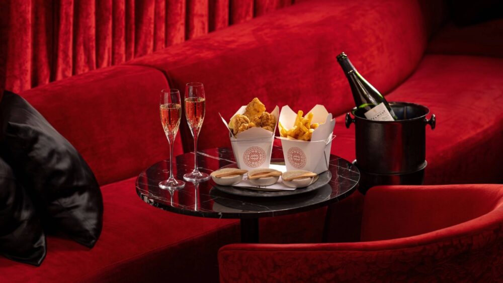 A black marble table set against red velvet seating holds champagne glasses, a bottle in a black ice bucket, and takeout boxes of fried chicken and truffle fries inside The Red Room at The Tampa EDITION.