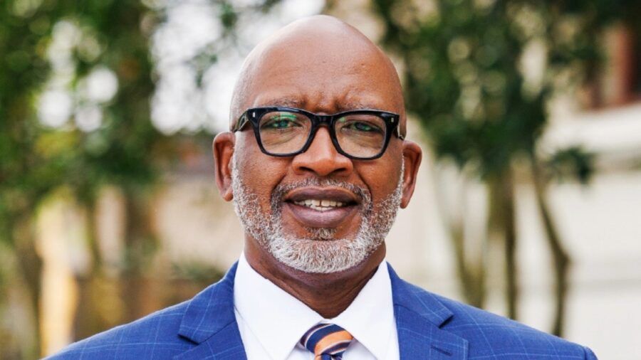 St. Petersburg Mayor Ken Welch wearing a blue suit and striped tie, standing outdoors with greenery in the background.