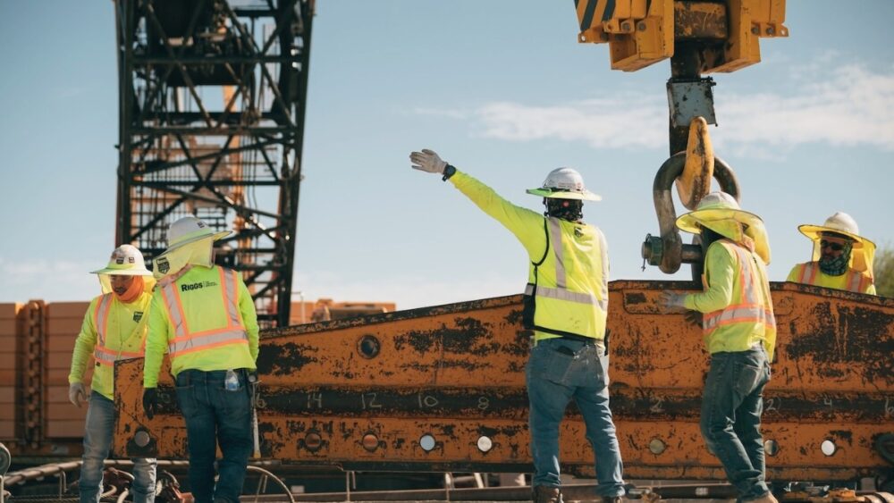 Construction workers in safety vests and hard hats guide heavy equipment at a job site, representing LGE Design Build’s on-site operations.