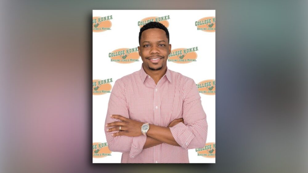 Roman Cowan standing with arms crossed in front of a brick wall, wearing a green College HUNKS uniform shirt.