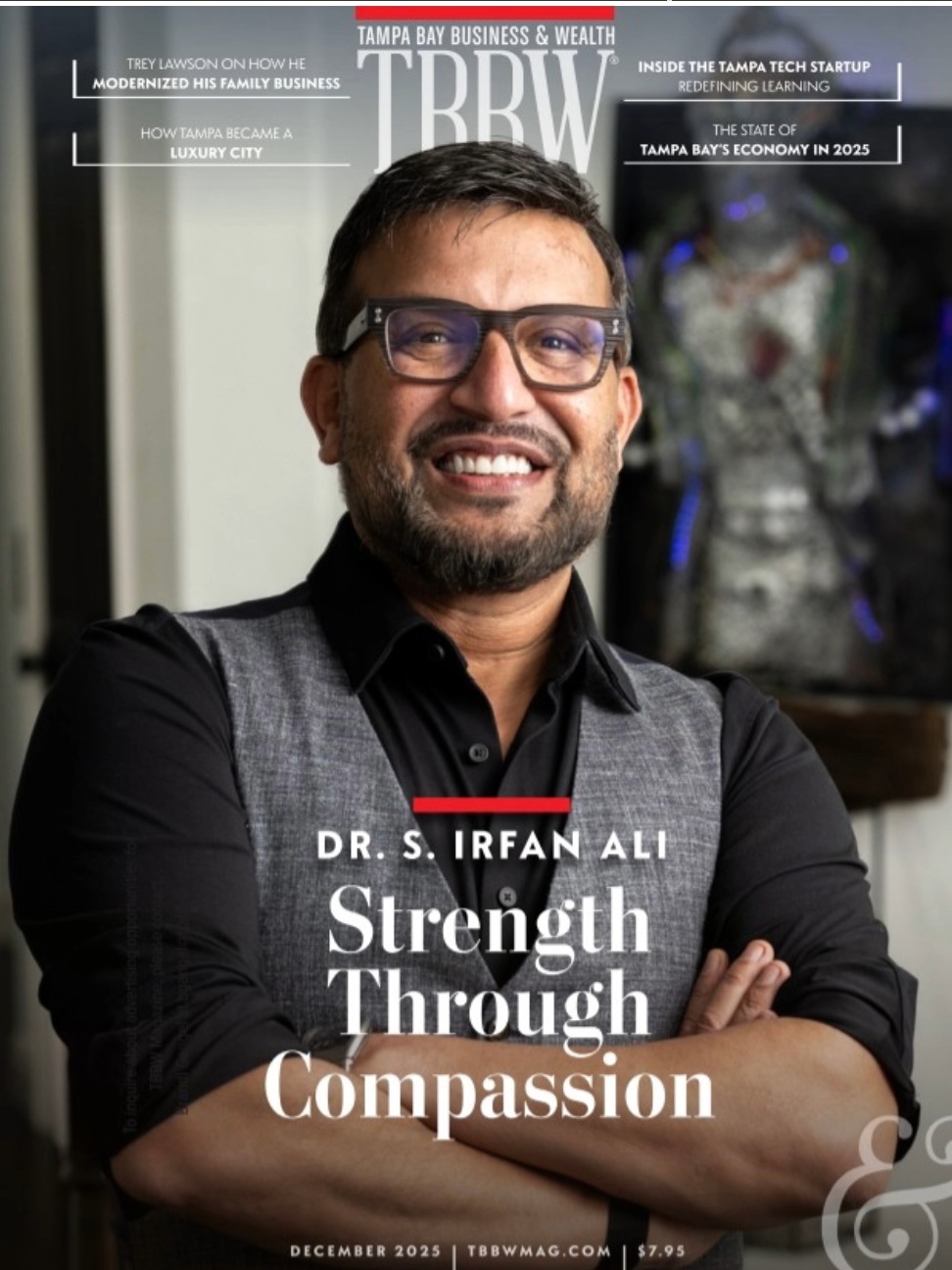 Dr. S. Irfan Ali stands with his arms crossed, smiling in front of artwork in his Tampa office. He is wearing glasses, a black shirt and a gray vest. The Tampa Bay Business and Wealth December 2025 cover headline reads “Strength Through Compassion.”