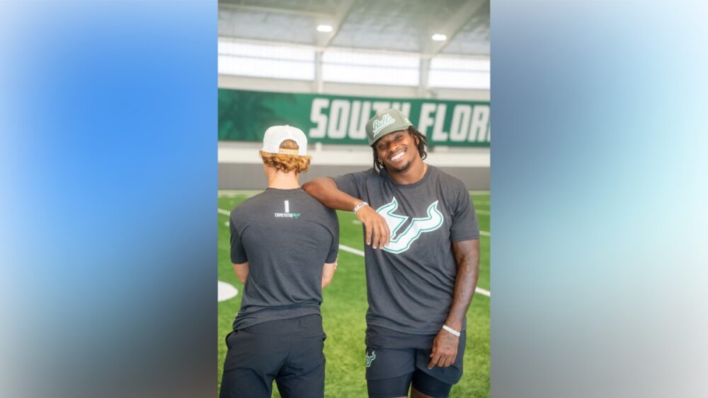 Two USF athletes wearing For the Bay shirts and hats on the indoor practice field at the University of South Florida