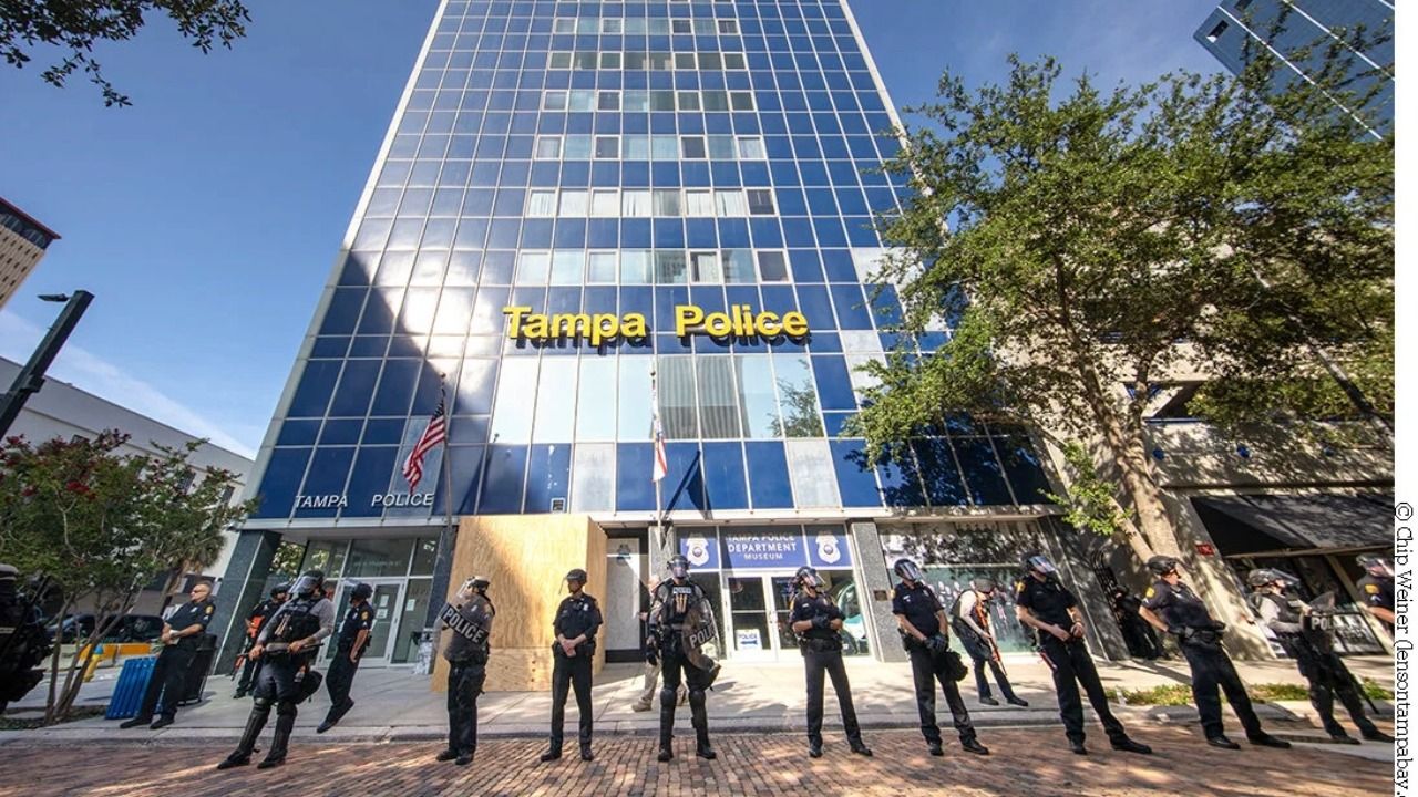 Tampa Police officers standing in a line in front of the Tampa Police Department headquarters, a tall blue-glass building in downtown Tampa.