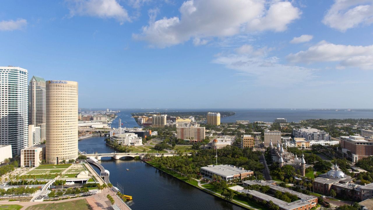 Aerial view of downtown Tampa showing the Hillsborough River, Riverwalk, high-rise office towers and neighborhoods stretching toward Tampa Bay under a bright blue sky.