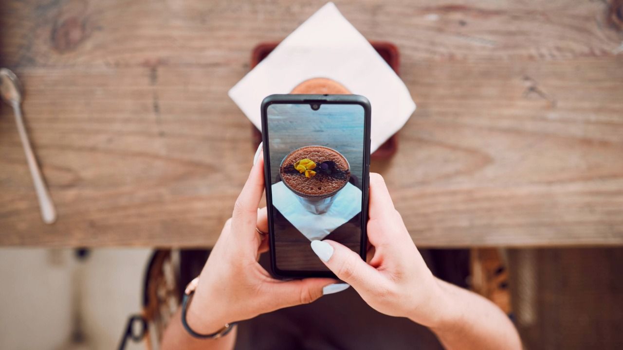 A person photographs a plated dessert with a smartphone at a café table, illustrating how food content is created for social media.