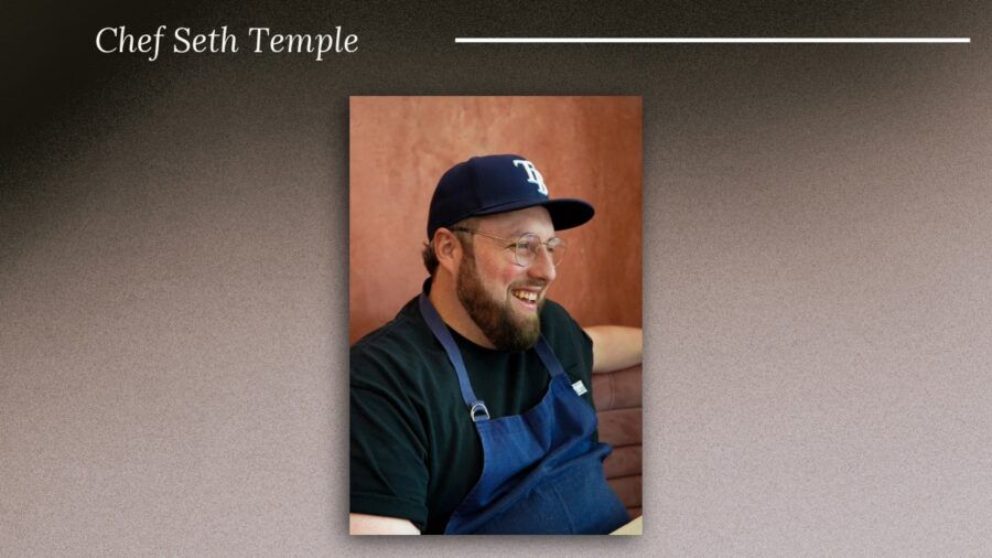 Chef Seth Temple smiles while seated inside ASH on Water Street, wearing a blue apron and Tampa Bay Rays cap.