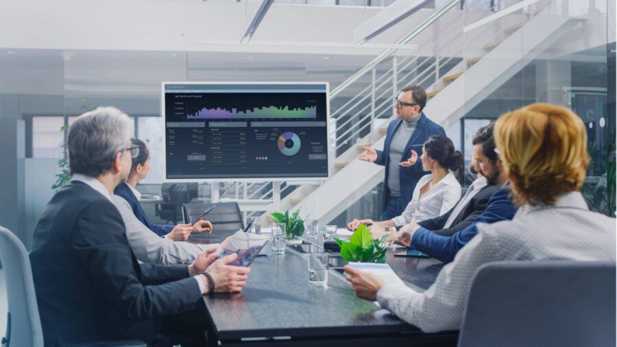 A business team meets in a modern conference room as a presenter points to a screen showing financial charts and market data.
