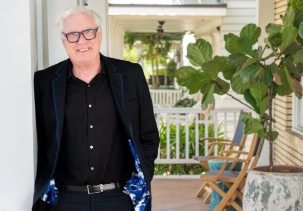 Bob Glaser stands on a covered porch in Tampa, wearing a black suit and smiling beside a potted plant.