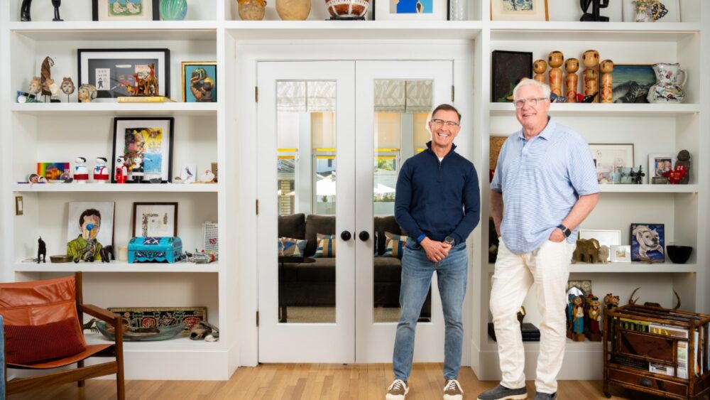 Bob Glaser and his husband, Miles, stand inside their home in front of built-in shelves filled with art and personal objects.
