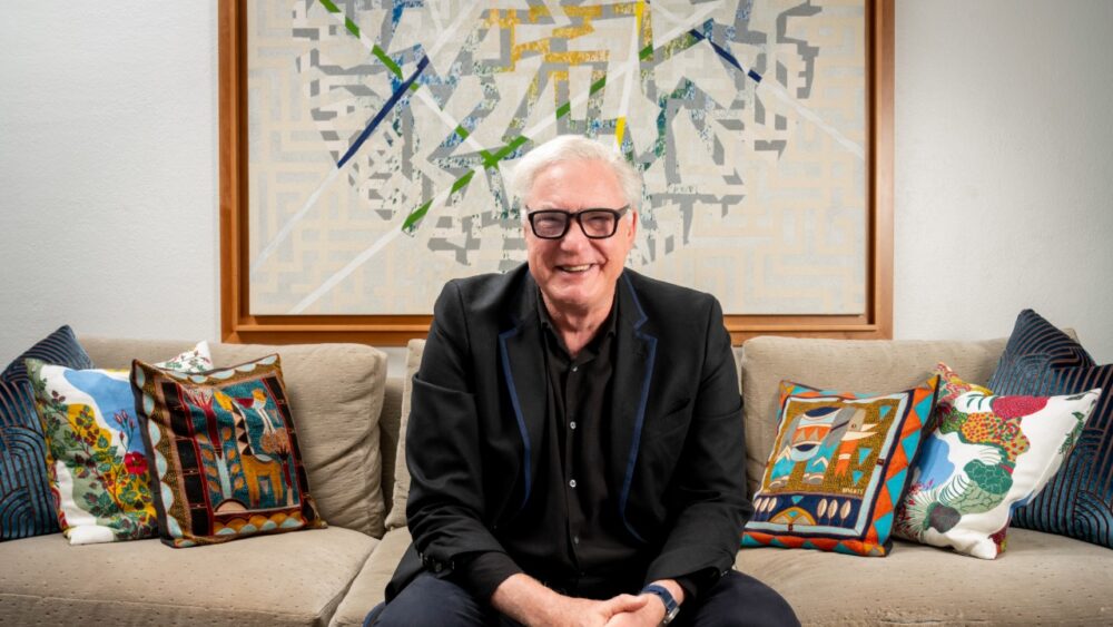 Bob Glaser sits on a couch in his home, framed by colorful pillows and a large abstract artwork behind him.