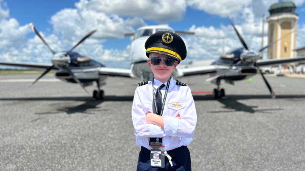 Child dressed as a pilot stands in front of an airplane on a runway