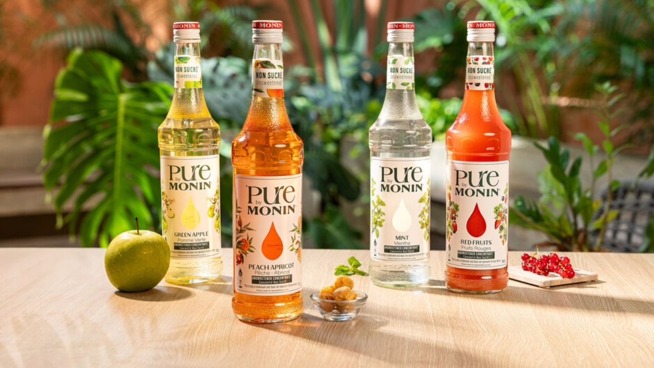 Four bottles of Pure by Monin beverage flavorings, including green apple, peach apricot, mint and red fruits, displayed on a wooden table with fresh fruit.
