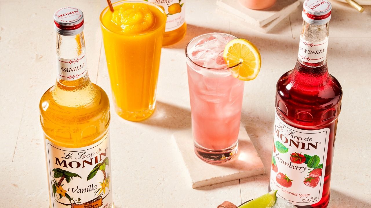Four bottles of Pure by Monin beverage flavorings, including green apple, peach apricot, mint and red fruits, displayed on a wooden table with fresh fruit.