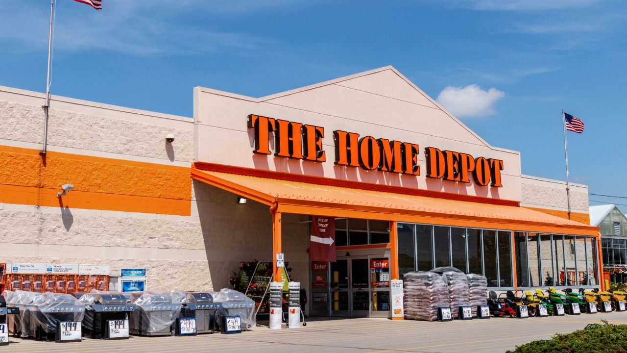 Exterior of a Home Depot store with merchandise displayed near the entrance and American flags overhead.