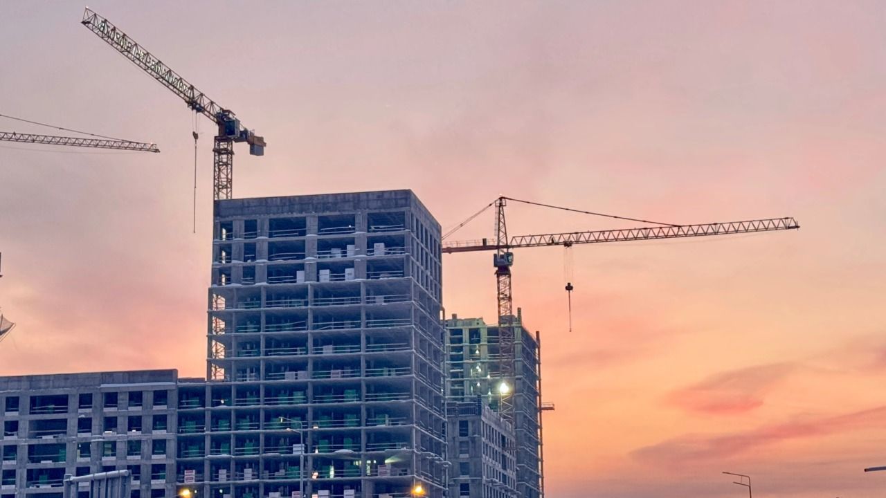 Construction cranes rise above a multistory building under construction against a pink sunset sky.