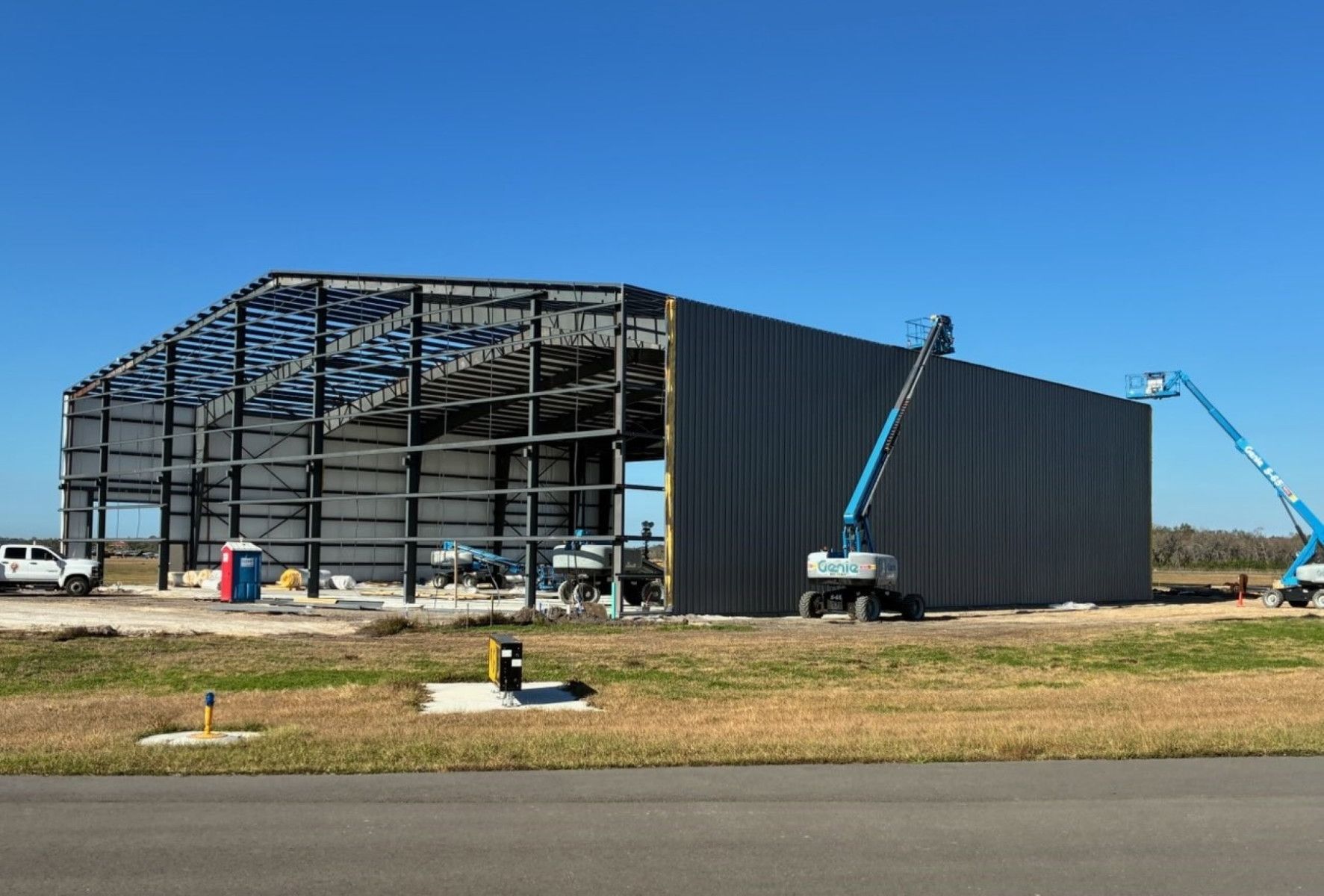 Large steel-frame aircraft hangar under construction, with cranes and lifts working on the exterior
