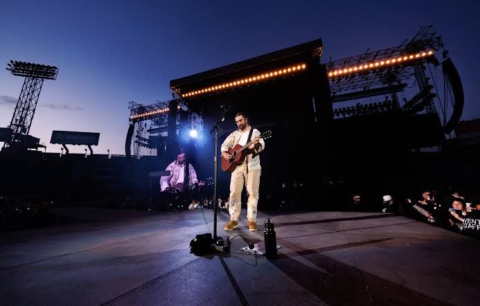 Noah Kahan performs with an acoustic guitar on an outdoor stadium stage at dusk.
