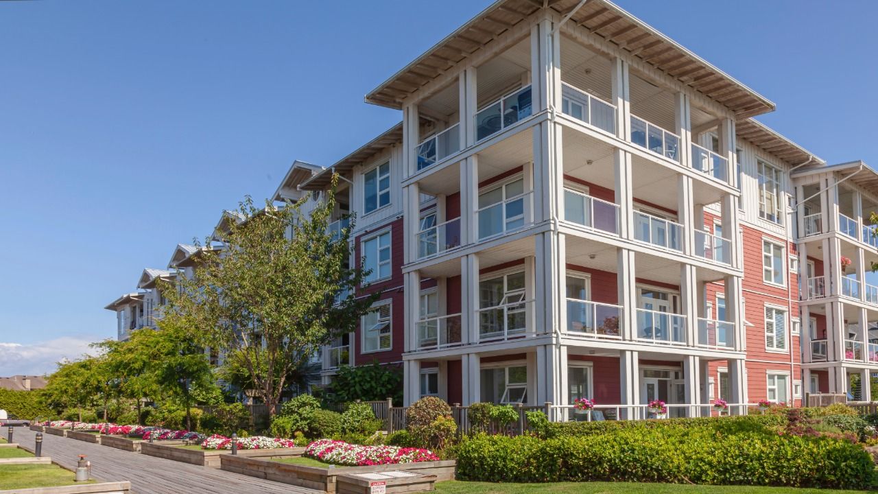 Four-story residential building with balconies, landscaped gardens and flower beds outside a retirement community.